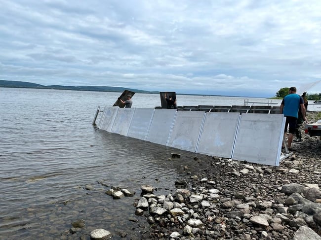 Three people assemble a portable, modular silver flood barrier along a rocky lake shoreline under a cloudy sky. The barrier consists of interconnected metal panels being positioned directly in the shallow water to prevent flooding before water levels rise.