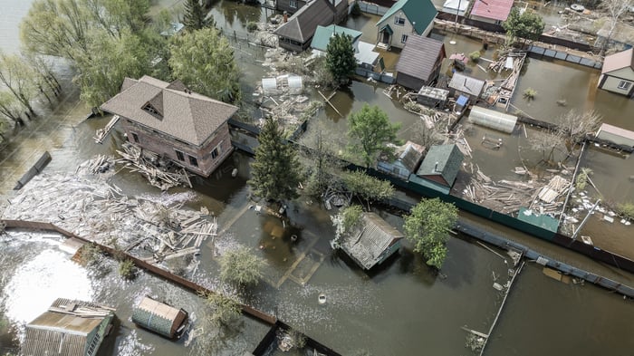 An aerial view shows a neighborhood submerged in brown floodwater. Several houses, surrounded by partially submerged fences and scattered wooden debris, stand in stagnant water that has overtaken yards and pathways. Green trees rise above the flooded landscape under bright daylight.